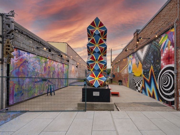 A colorful geometric sculpture stands in an outdoor art alley, surrounded by vibrant murals on brick walls and string lights overhead, with a dramatic sunset sky in the background.
