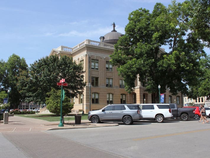 A historic courthouse with a domed roof stands behind trees and several parked cars on a sunny day. A red street sign and people walking are visible in the foreground.