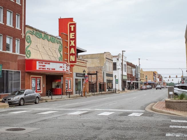 A small-town Main Street with historic buildings, including a red and yellow “TEXAN” theater marquee, parked cars, and empty sidewalks under an overcast sky.
