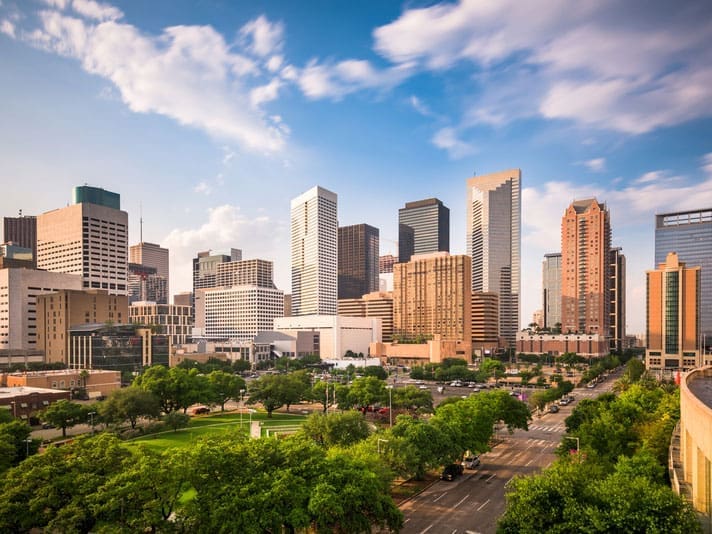 Downtown cityscape with modern high-rise buildings, a blue sky with scattered clouds, green trees, and a park in the foreground beside a wide street with cars.