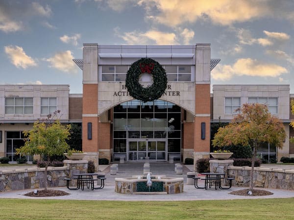 A modern activity center with large glass doors, a wreath above the entrance, and a small fountain surrounded by picnic tables in front. Trees with autumn foliage line the walkway. The sky is partly cloudy.