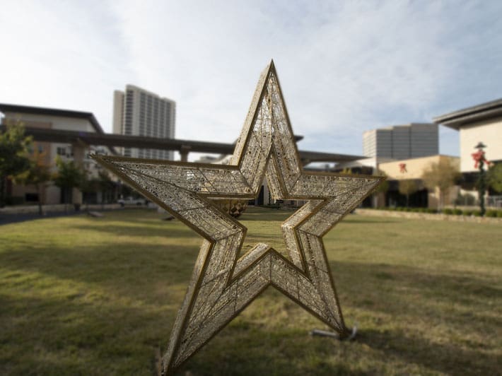 A large decorative star sculpture stands on a grassy lawn with tall buildings and trees in the background under a partly cloudy sky.