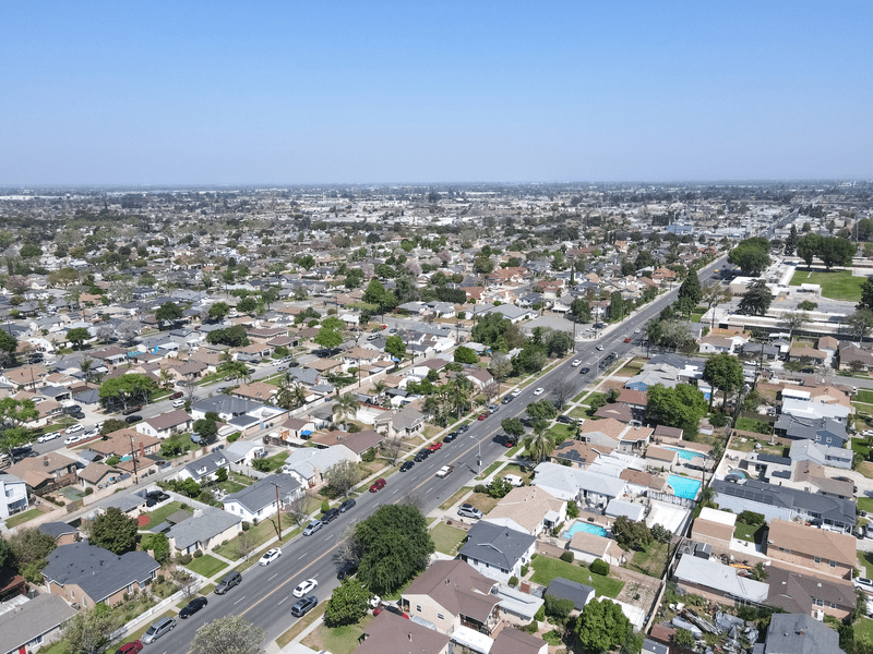 Aerial view of a suburban neighborhood with a wide road running through the center, lined with houses, trees, and cars, stretching into the distance under a clear blue sky.