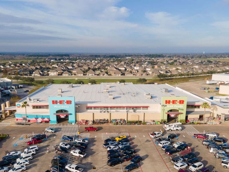 Aerial view of an H-E-B supermarket with a busy parking lot full of cars in front. The store has colorful entrances, and residential neighborhoods can be seen in the background under a blue sky.