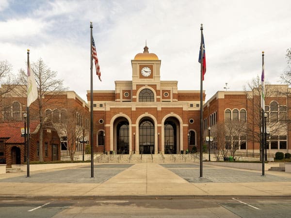 A large brick building with a central clock tower, arched entrance, and multiple flagpoles in front. Leafless trees and paved walkways are visible, suggesting it is fall or winter.