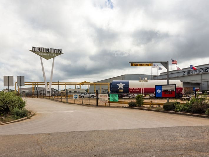 Industrial facility with a large Trinity Industries sign on tall supports, trucks parked outside, a fenced entrance, and flags flying. Overcast sky with thick clouds above the building.