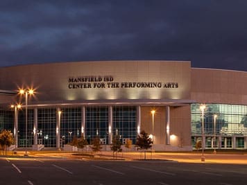 Exterior view of the Mansfield ISD Center for the Performing Arts at dusk, with illuminated windows and an empty parking lot in front. The building name is visible on the facade.