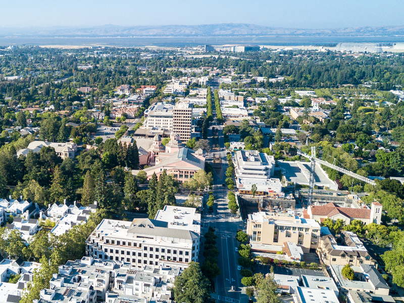Aerial view of a city with tree-lined streets, various buildings, and distant mountains under a clear blue sky.