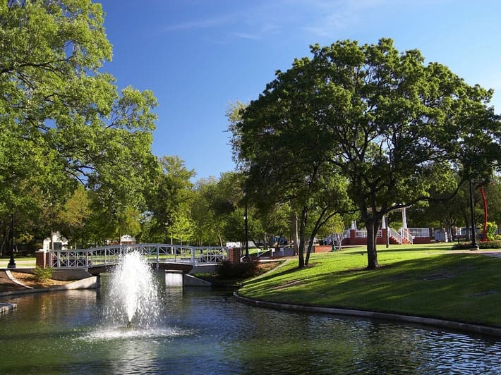 A sunny park scene with a small pond featuring a water fountain, a white footbridge, lush green grass, and large leafy trees under a clear blue sky.