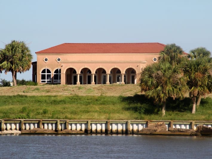 A large, tan stucco building with a red tile roof and arched windows sits on a grassy hill near palm trees, overlooking a body of water with a reinforced shoreline.