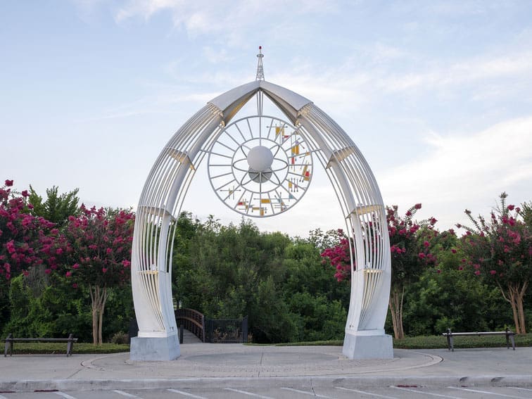 A white metal arch sculpture with a circular, abstract design at the top stands in front of green trees and pink flowering bushes. Two park benches sit nearby under a blue sky with wispy clouds.