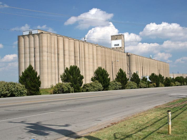 Large concrete grain elevator with multiple silos stands beside a wide, empty road. Green trees and bushes line the building, and a blue sky with scattered clouds is visible above.