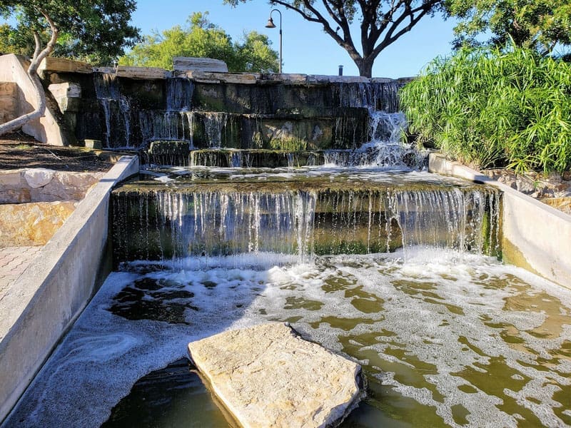 A small multi-tiered stone waterfall flows into a shallow pool surrounded by greenery and trees under a clear blue sky. A flat rock sits in the water at the front of the image.