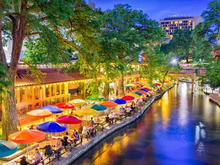 A lively riverside scene at dusk with colorful umbrellas shading outdoor dining tables along a tree-lined waterway in a city, illuminated by warm lights from nearby buildings.