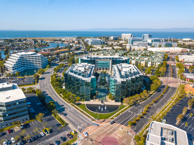 Aerial view of modern office buildings with glass facades, arranged in a circular pattern near a main road, surrounded by parking lots, trees, and other commercial buildings, with water and cityscape in the background.