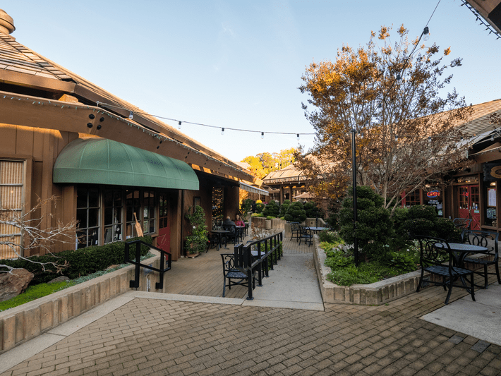 Outdoor courtyard with tables and chairs surrounded by shops and greenery, string lights overhead, trees with autumn leaves, and buildings with wooden exteriors under clear blue sky.