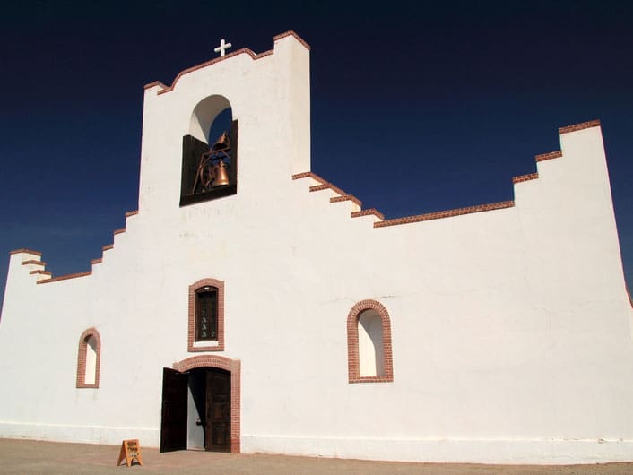 A white adobe church with a stepped roofline, arched windows, a bell tower topped with a cross, and a large wooden door, set against a clear blue sky.