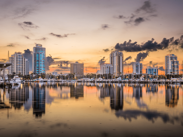 A city skyline with tall buildings and palm trees is reflected in a calm marina at sunset, with boats docked in the harbor and clouds scattered across a colorful sky.
