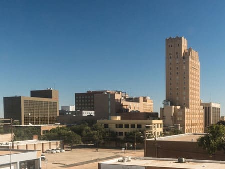 A cityscape featuring several mid-rise buildings and a prominent tall, tan, art deco-style tower against a clear blue sky, with a few trees and streets in the foreground.