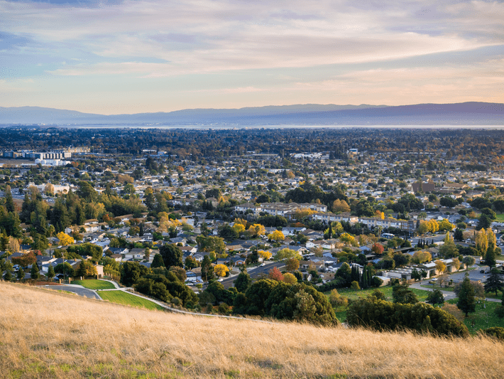 A panoramic view of a suburban city with houses and trees, seen from a grassy hillside under a partly cloudy sky, with distant mountains in the background.