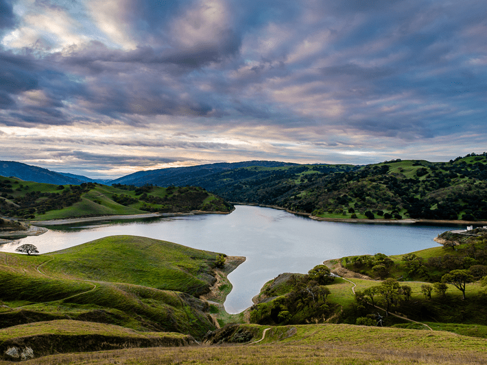A serene lake surrounded by rolling green hills under a dramatic, cloudy sky at sunset, with winding paths visible along the landscape.