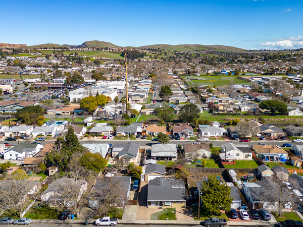 Aerial view of a suburban neighborhood with rows of houses, green lawns, and tree-lined streets, set against rolling hills and a clear blue sky in the background.