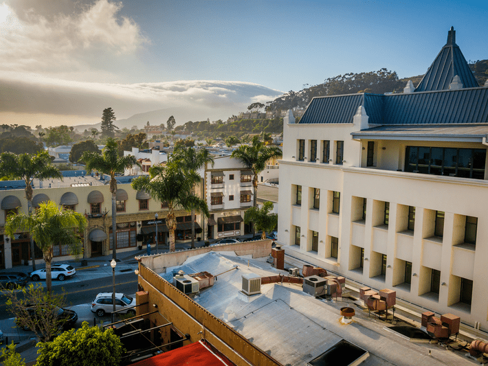A cityscape featuring palm trees, modern and Mediterranean-style buildings, and rolling hills in the background under a partly cloudy sky at sunset.