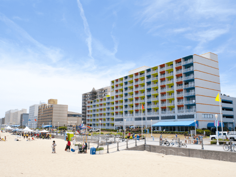 A sunny beach scene with people relaxing on the sand, a row of mid-rise beachfront hotels, bicycles parked by a railing, and a blue sky with wispy clouds overhead.