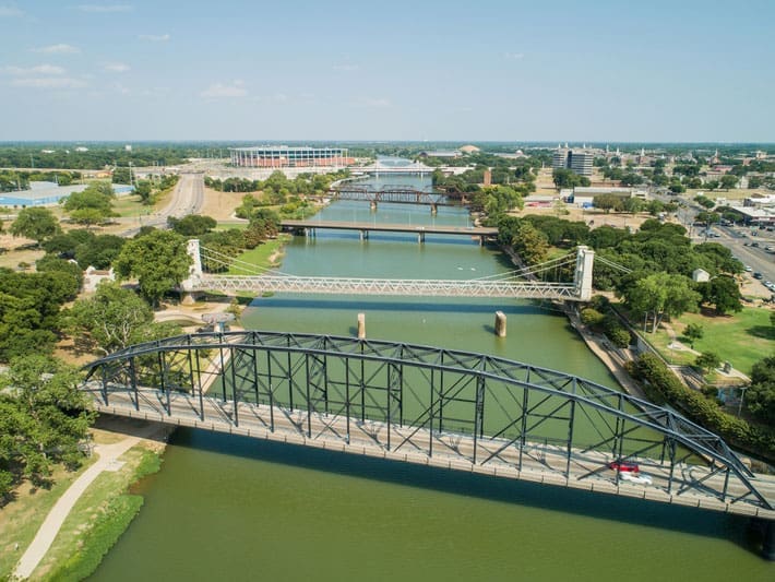 Aerial view of multiple bridges spanning a wide river with green banks, trees, and nearby roads; buildings and a stadium are visible in the background under a clear sky.
