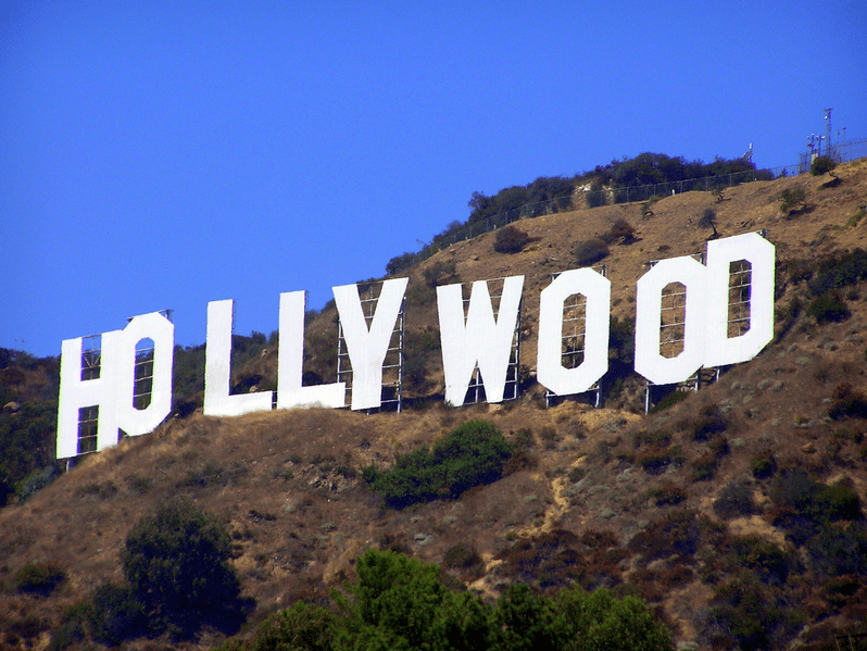 Large white HOLLYWOOD sign stands on a dry, shrub-covered hillside under a clear blue sky, surrounded by scattered bushes and trees.