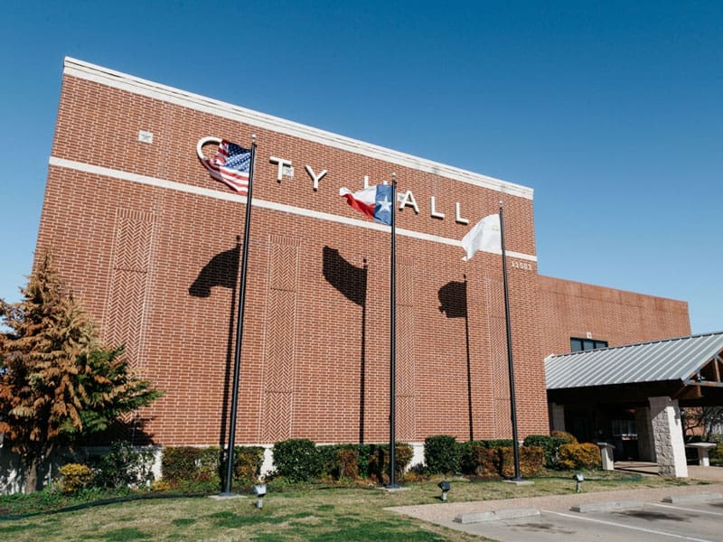 A red brick building labeled CITY HALL with three flagpoles in front, displaying the U.S., Texas, and a white flag. Shadows of the flags are cast on the building. There is a clear blue sky above.