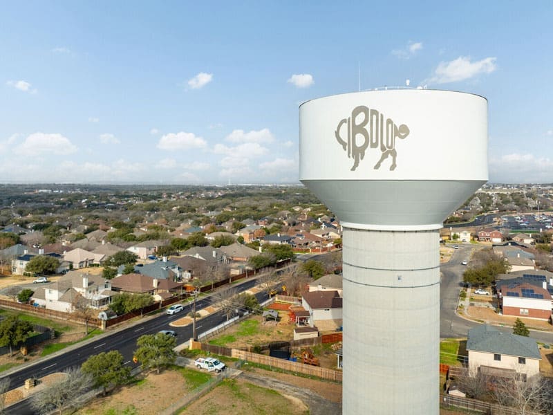 A tall water tower with a simple buffalo logo stands over a suburban neighborhood with houses, trees, and roads under a partly cloudy sky.