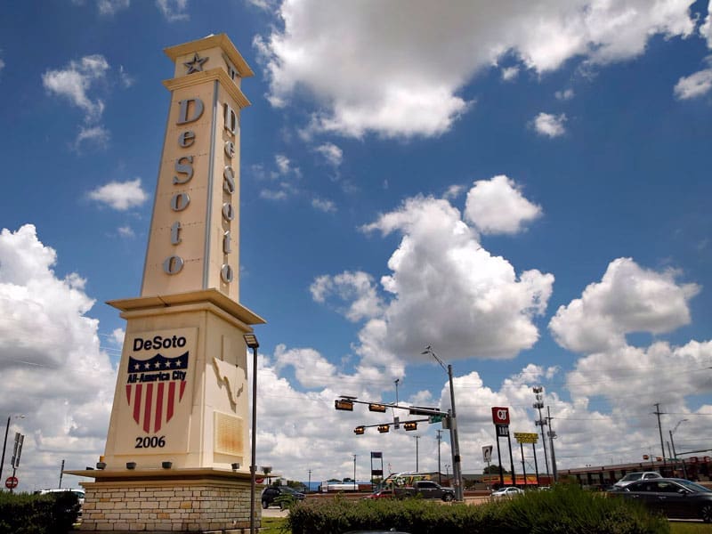 A large pillar sign reads DeSoto, All-America City, 2006 against a blue sky with scattered clouds. Traffic lights, street signs, and businesses are visible in the background.