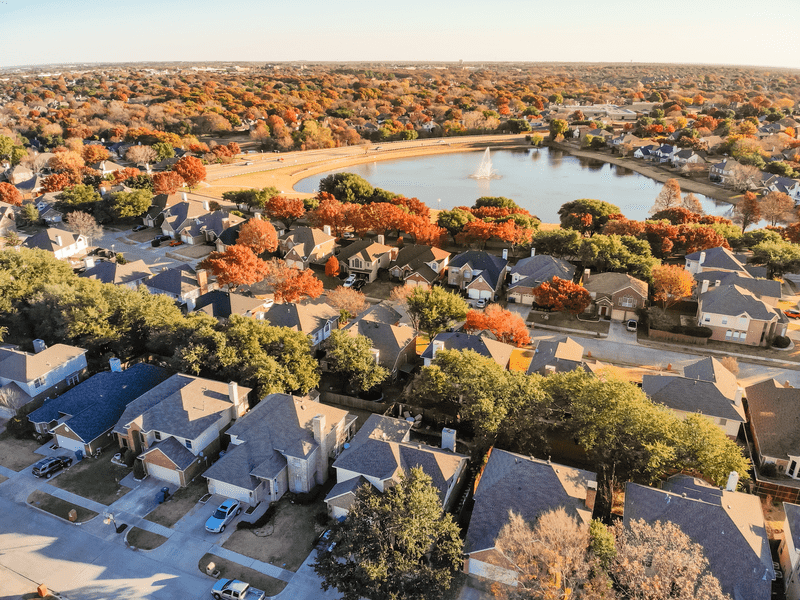 Aerial view of a suburban neighborhood with rows of houses, tree-lined streets displaying autumn colors, and a small lake with a fountain at the center, surrounded by more homes and trees.