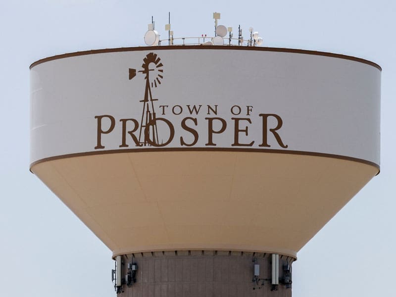 A large white water tower with Town of Prosper and a windmill graphic painted on it, set against a light sky. Several antennas and equipment are mounted on top.